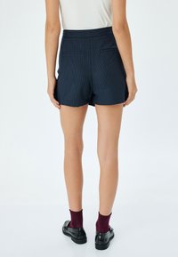 Marineblauwe korte broek van een textuurstof, met een hoge taille, twee achterzakken en een getailleerde pasvorm. Draagt met zwarte schoenen en bordeauxrode sokken.