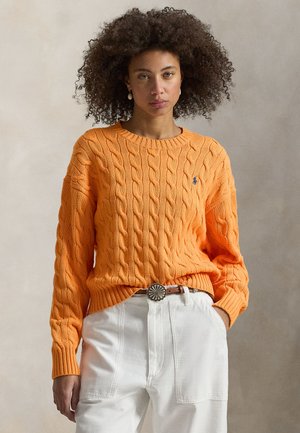 Orange cable-knit sweater with ribbed cuffs and hem, paired with white high-waisted pants and a decorative belt buckle.