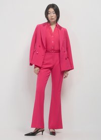 Costume ajusté rose vif avec un blazer à double boutonnage et un pantalon évasé. Le blazer a de grands boutons et des revers en satin. Chaussures noires à talons.