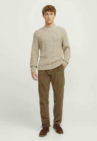 Jeune homme debout, portant un pull en tricot beige, un pantalon en velours côtelé marron et des chaussures à lacets marron, devant un fond clair et uni.