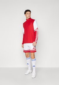 Punane ja valge sportlik riietus, millel on pikkade varrukatega tõmbluku top, triibulised lühikesed püksid ja valged saapad siniste aktsentidega.