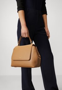 Kabelka z tanového kůže se strukturovaným tvarem, horní rukojetí, zlatými kovovými detaily a klopovým zapínáním. Hladký povrch, minimální značení.