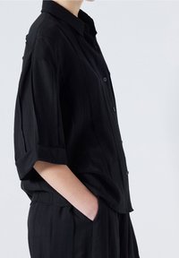 Chemise noire à boutons avec manches retroussées, en tissu lisse présentant de fines lignes verticales. Poche latérale visible sur la hanche du mannequin.