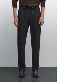 Pantaloni neri con vestibilità rilassata, vita con coulisse e tessuto liscio. Il modello indossa scarpe scure e sta in piedi su un pavimento scuro a macchie.