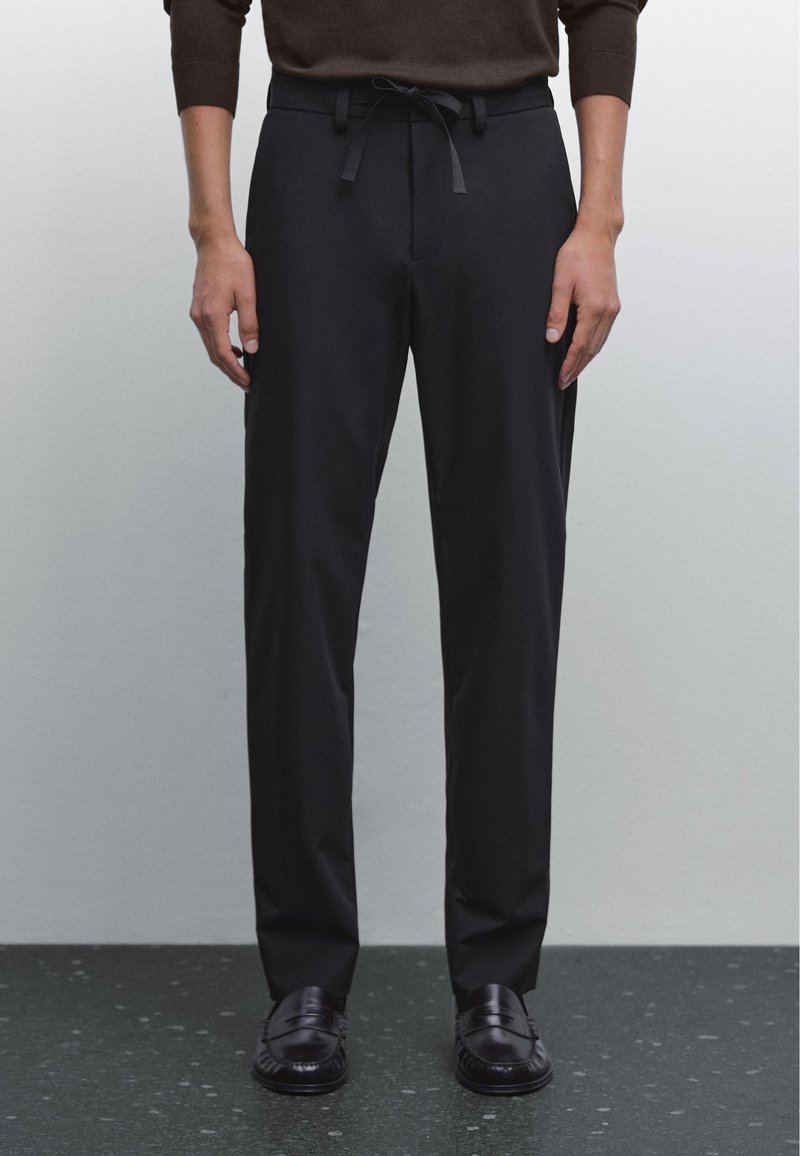 Pantaloni neri con vestibilità rilassata, vita con coulisse e tessuto liscio. Il modello indossa scarpe scure e sta in piedi su un pavimento scuro a macchie.