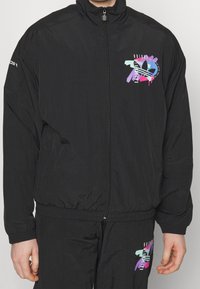 Svart vindjacka i lättviktstyg. Har en färgstark grafisk tryck på vänstra bröstet samt elastisk midja och muddar.
