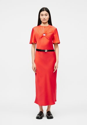 Γυναίκα με μακριά σκούρα μαλλιά φοράει midi φόρεμα από κόκκινο σατέν με κεντρικό στρογγυλό άνοιγμα και μαύρη ζώνη, συνδυασμένο με μαύρα μοκασίνια.