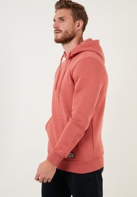 Sudadera con capucha rosa, con un bolsillo frontal, puños y dobladillo acanalados. Presenta cordones y una etiqueta con logo en la parte inferior. Textura suave.