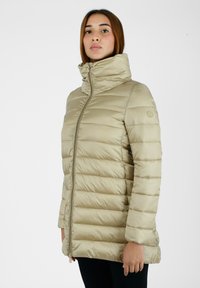 Giacca imbottita beige, morbida, con colletto alto, cuciture orizzontali, zip frontale, tasche laterali e finitura liscia e lucida.