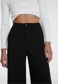 Zwarte wijde broek met een knoopsluiting aan de voorkant, vlakke tailleband en een gladde textuur, gecombineerd met een witte geribde top met lange mouwen.