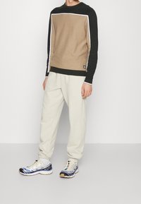 Felpa a maniche lunghe marrone e nera con un pannello rettangolare beige; abbinata a pantaloni sportivi crema e sneakers bianche con accenti blu.