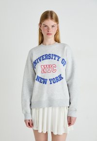 Jovem mulher com cabelo loiro longo, vestindo um moletom cinza com o texto "Universidade de Nova Iorque NYC" e uma saia branca, posando contra um fundo simples.