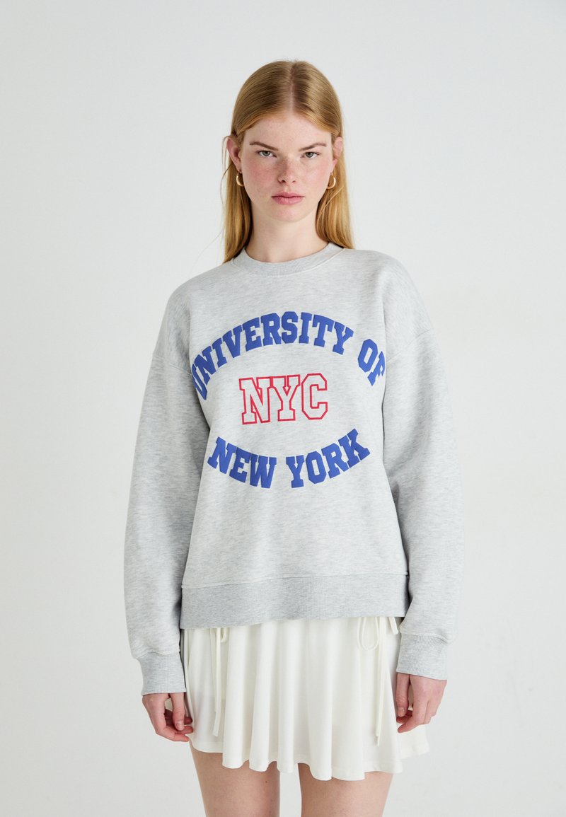 Jovem mulher com cabelo loiro longo, vestindo um moletom cinza com o texto "Universidade de Nova Iorque NYC" e uma saia branca, posando contra um fundo simples.