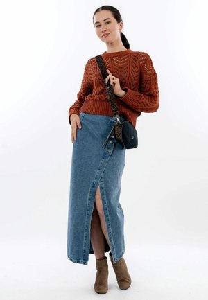 Maglione lavorato a maglia ruggine con motivo a trama aperta, abbinato a una maxi gonna in denim blu con spacco laterale e dettagli di bottoni, e stivaletti taupe alla caviglia.