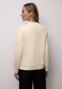 Sweatshirt à enfiler de couleur crème avec un col montant, des poignets et un ourlet côtelés. Texture lisse avec une coupe décontractée et un détail de couture centrale à l'arrière.
