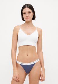 Bralette en coton blanc avec des bretelles fines, associé à un string bleu avec une ceinture blanche Calvin Klein. Texture lisse, design minimaliste.