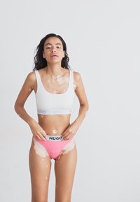 Hvit sport bh med blå "HUGO" midjebånd og rosa bikini truser med svart "HUGO" midjebånd; glatt stoff, tettsittende stil, minimalistisk design.