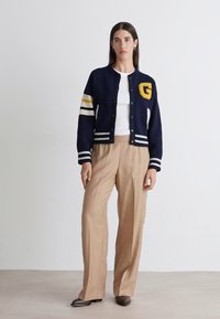 Ναυτικό varsity μπουφάν με κίτρινο λεπτομέρεια "G", ριγέ μανίκια και λευκές λεπτομέρειες, συνδυασμένο με μπεζ φαρδιά παντελόνια και λευκό μπλουζάκι.