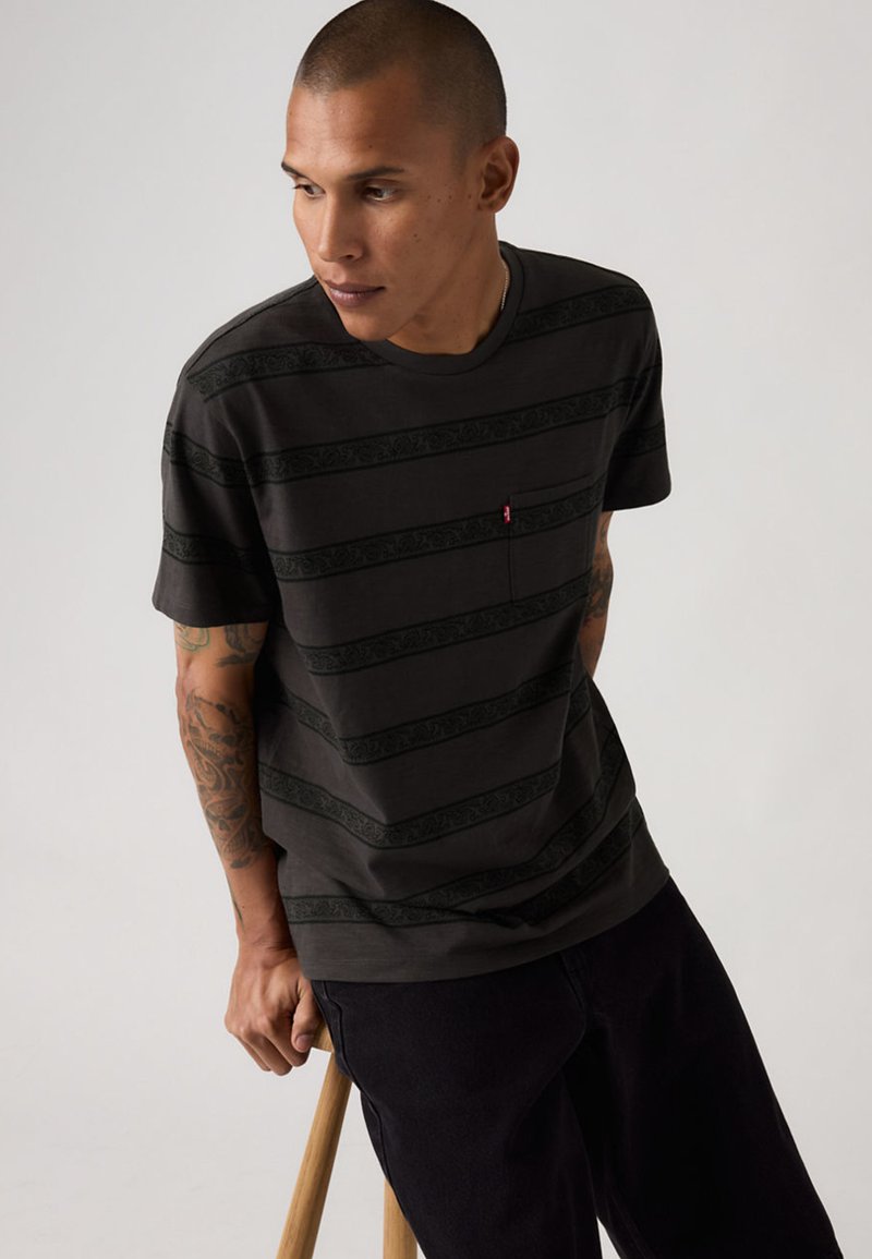 Homme tatoué assis sur un tabouret en bois, portant une chemise à manches courtes noire rayée et un pantalon sombre, regardant sur le côté sur un fond uni.