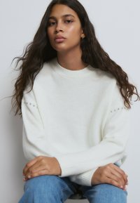 Junge Frau mit langen, welligen Haaren, die einen weißen Pullover mit Perlendetails an den Ärmeln trägt und mit den Händen auf blauer Jeans sitzt.