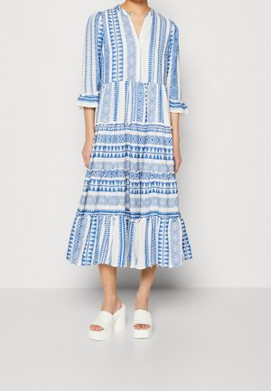 Biała sukienka midi z niebieskimi geometrycznymi wzorami, rękawy do łokcia, falujące warstwy, w połączeniu z białymi sandałami na wysokim obcasie na platformie.