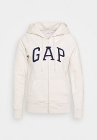 Sudadera con cremallera de color crema claro, hecha de una tela suave. Presenta grandes letras en azul marino con la palabra "GAP" en el frente y bolsillos tipo canguro. Diseño acogedor.