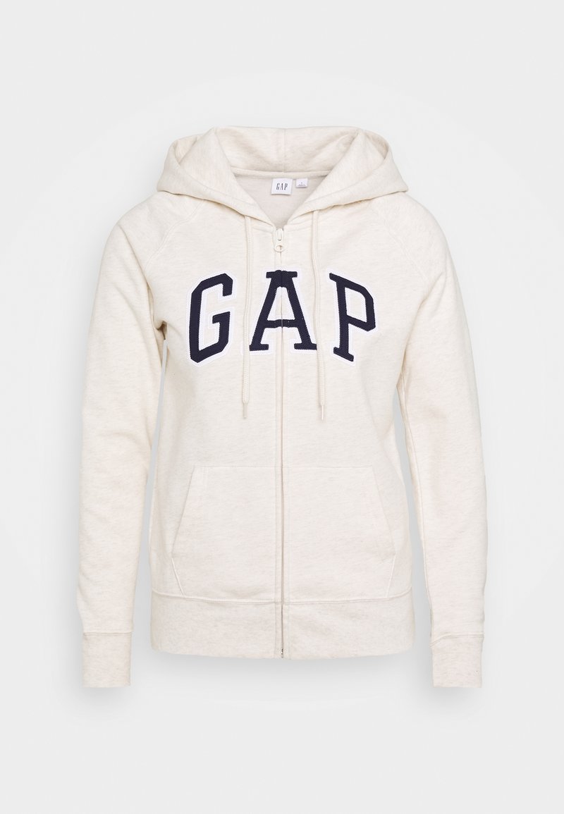 Sudadera con cremallera de color crema claro, hecha de una tela suave. Presenta grandes letras en azul marino con la palabra "GAP" en el frente y bolsillos tipo canguro. Diseño acogedor.