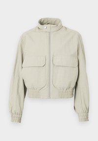 Veste cropped gris clair avec un col montant, fermeture éclair devant et deux grandes poches poitrine. Les poignets et l'ourlet élastiqués ajoutent de la texture.