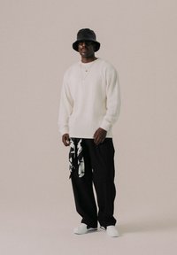 Suéter blanco de punto de manga larga, pantalones negros de pierna ancha con estampado floral, sombrero negro tipo bucket, gafas de sol negras, zapatos blancos, collares dorados en capas.