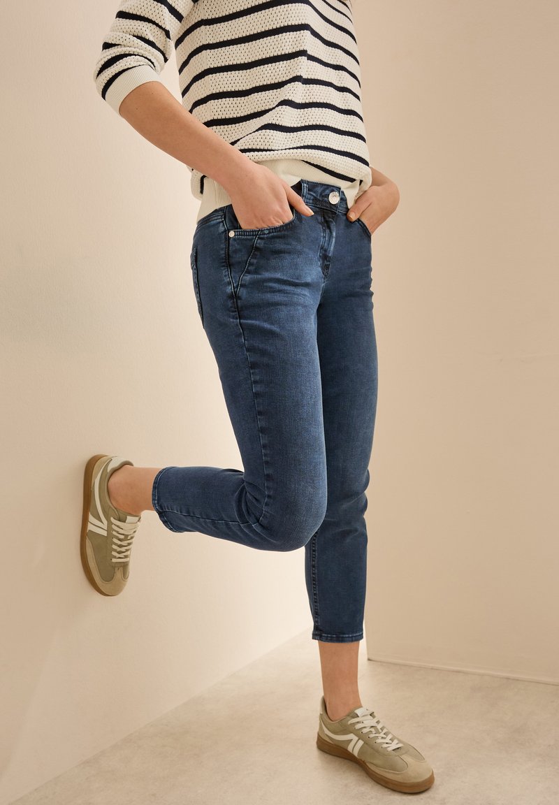 Jean en denim bleu avec une coupe slim, design à cinq poches et taille mi-haute. Assortis avec des baskets beiges et blanches. Pull rayé visible.