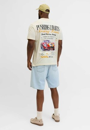 Hombre con gorra beige, camiseta color crema con gráfico de coche de carreras rojo y texto, pantalones cortos de mezclilla claros, zapatos beige, de pie sobre fondo blanco.