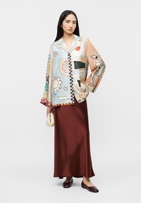 Γυναίκα με πολύχρωμο σακάκι με αφηρημένο σχέδιο, μακριά καφέ φούστα και σκούρα καφέ μπαλαρίνες, κρατώντας μία μικρή μπεζ τσάντα.