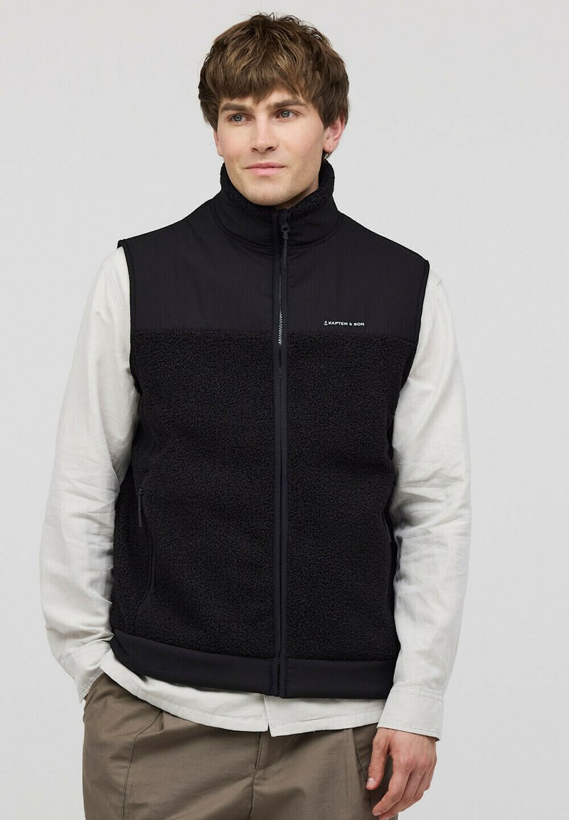 Schwarze Fleece-Weste mit glattem Oberteil und strukturiertem Unterteil, hohem Kragen und seitlichen Reißverschlusstaschen. Logo auf der Brust.