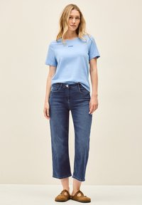 Camiseta de algodón azul claro con un pequeño logotipo bordado, combinada con jeans recortados azul oscuro y zapatos de ante marrón sin cordones.