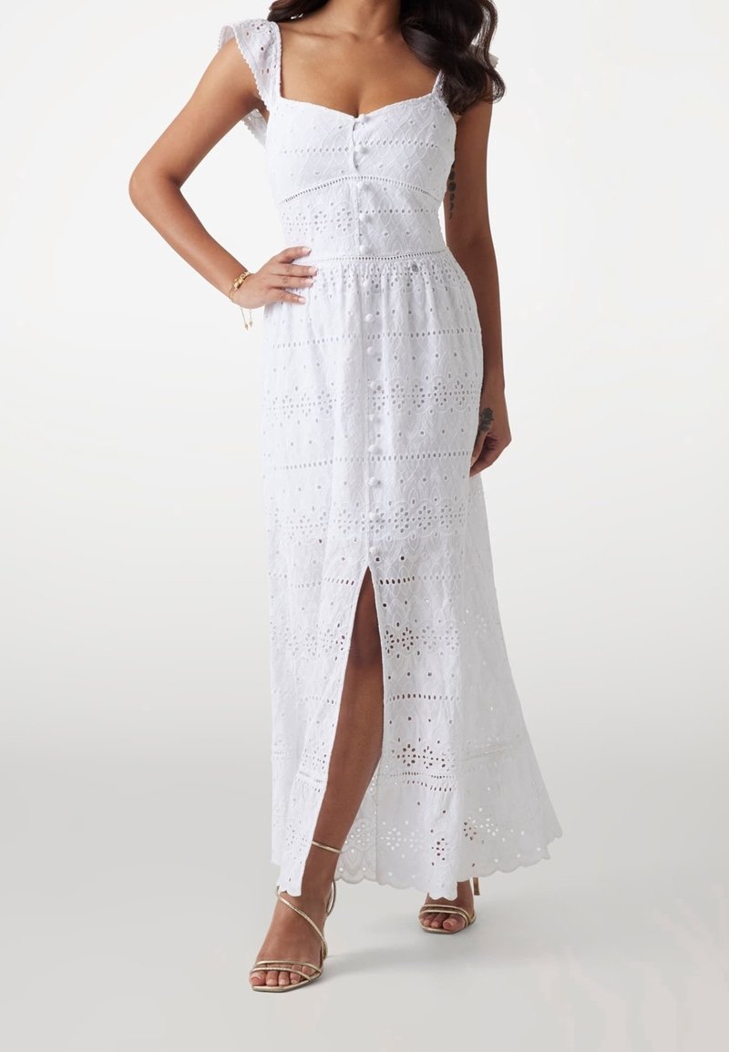 Femme portant une robe maxi blanche à œillets avec des boutons sur le devant et une fente devant, associée à des sandales à brides dorées.