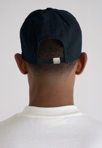 Persona che indossa un cappellino da baseball nero al contrario con cinturino regolabile, con una treccia visibile, e indossa una maglietta bianca a girocollo, di spalle.