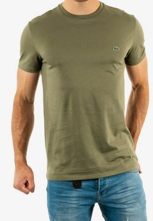 Homme portant un t-shirt à manches courtes vert olive avec un petit logo crocodile sur la poitrine et un jean bleu délavé, debout avec le poing serré.