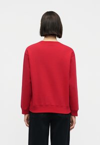 Άτομο με κοντά σκούρα μαλλιά, φορώντας ένα απλό κόκκινο φούτερ και μαύρο παντελόνι, εμφανιζόμενο από την πίσω πλευρά σε μία απλή λευκή φόντο.