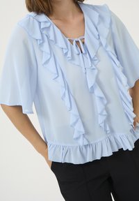 Jasnoniebieska bluzka z falbanami przy dekolcie i rękawach, z wiązaniem z przodu, wykonana z przezroczystej, miękkiej tkaniny.