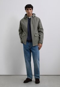 Chaqueta con capucha color caqui, con cremallera y dos bolsillos frontales, combinada con jeans azules y zapatos marrón oscuro. Textura suave, diseño casual.