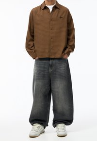 Camisa de cuadros marrón con botones, de corte relajado y un pequeño logo, combinada con vaqueros grises sueltos y zapatillas blancas.