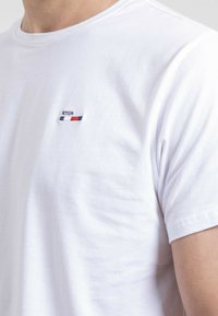 T-shirt en coton blanc avec col rond. Présente un petit logo brodé en marine, rouge et blanc sur la poitrine gauche. Texture douce.