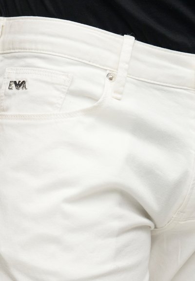 Κοντινή λήψη λευκών τζιν παντελονιών με ασημένιο καρφίτσα με το λογότυπο EVA στην μικρή μπροστινή τσέπη και μεταλλικό κουμπί πάνω από το φερμουάρ.