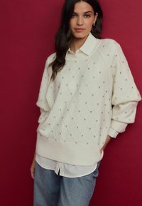 Pull crème avec un motif en losange, agrémenté de petites clous argentés, coupe oversize, tricot texturé, porté par-dessus une chemise blanche à col.