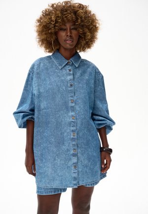 Kobieta z kręconymi włosami ubrana w oversize'ową niebieską sukienkę koszulową z denimu i duże kołowe kolczyki, stojąca na białym tle.