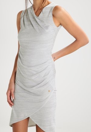 Ærmeløs lysegrå figursyet kjole med draperet front og asymmetrisk kant på en stående person med den ene hånd på hoften.