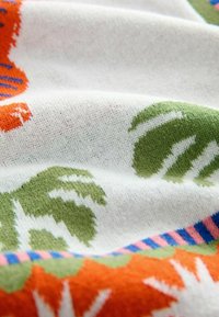 Měkká bílá látka s pletenými zelenými listy a oranžovými květinovými vzory, zobrazující texturu a barevné pruhované okraje.