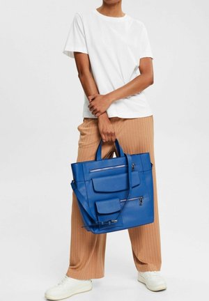 Sac fourre-tout bleu avec plusieurs poches avant, une surface texturée, des anses robustes, et une bandoulière amovible, accompagné de pantalons plissés beiges.