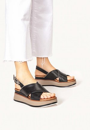Czarne skórzane sandały na koturnie z krzyżującymi się paskami, beżową platformą i białymi akcentami. Regulowany pasek wokół kostki z metalowymi elementami.