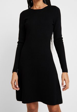 Woman wearing a fitted black long-sleeve dress with a round neckline, standing against a plain light background.